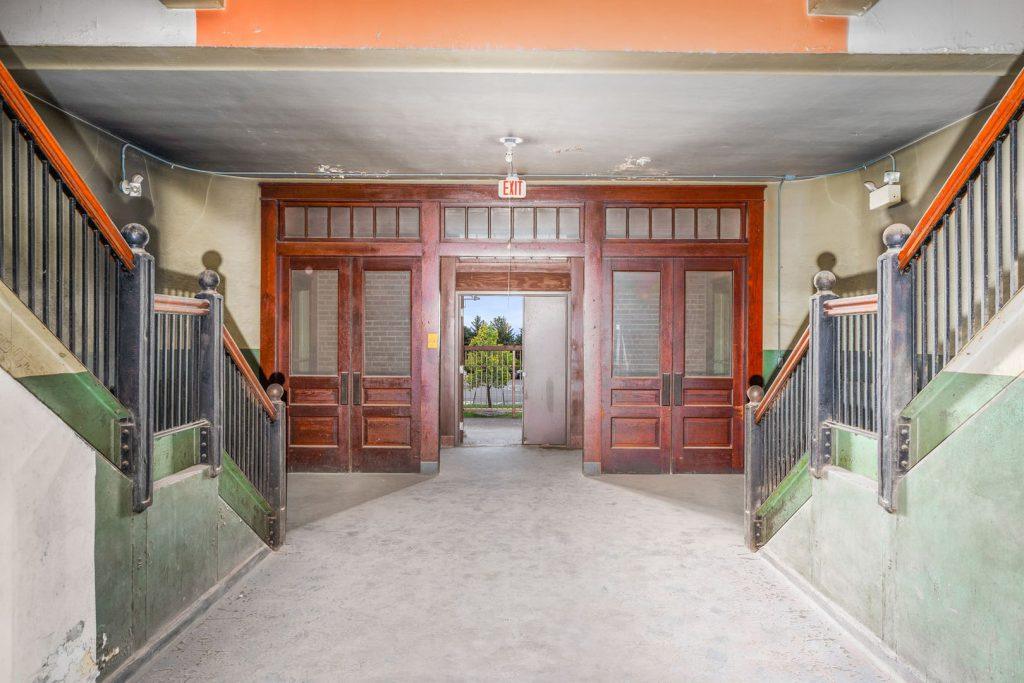 Old school hallway with double wooden doors and staircases on each side, leading to an exit with natural light and greenery visible outside. The walls and stairs show signs of wear.