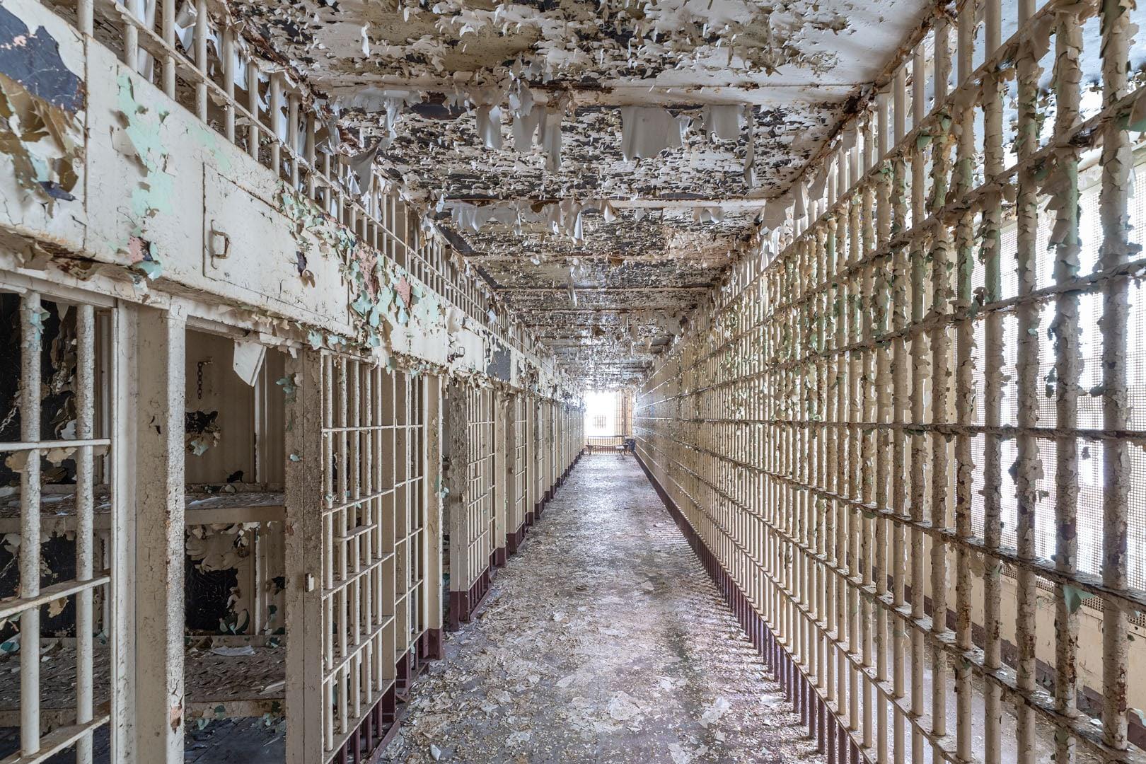 A long, abandoned prison corridor with peeling paint, rusted metal bars, and decaying walls, leading to a bright light at the far end. The scene looks weathered and neglected.