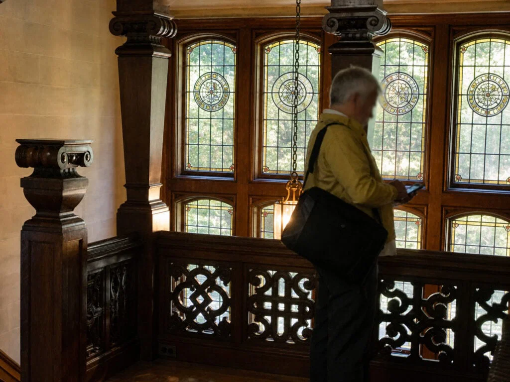 A person with a shoulder bag stands near a wooden railing inside a building with large stained glass windows, partially illuminated by sunlight.