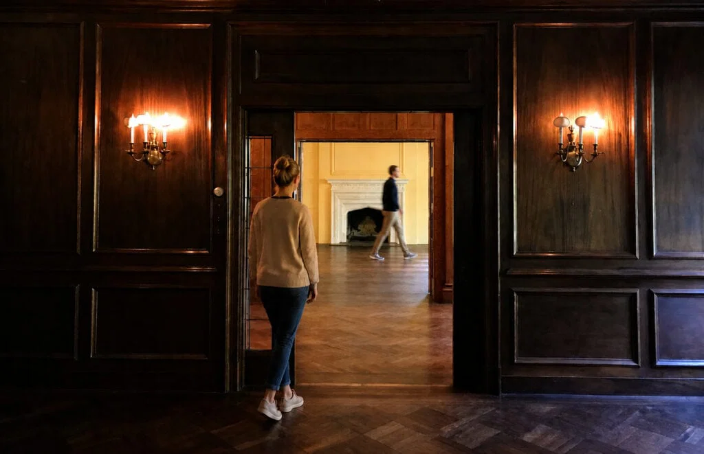 A woman stands in a dim, wood-paneled room, looking through a doorway into a brighter room where a man walks past a white fireplace. Two wall sconces with lit bulbs are on either side of the doorway.