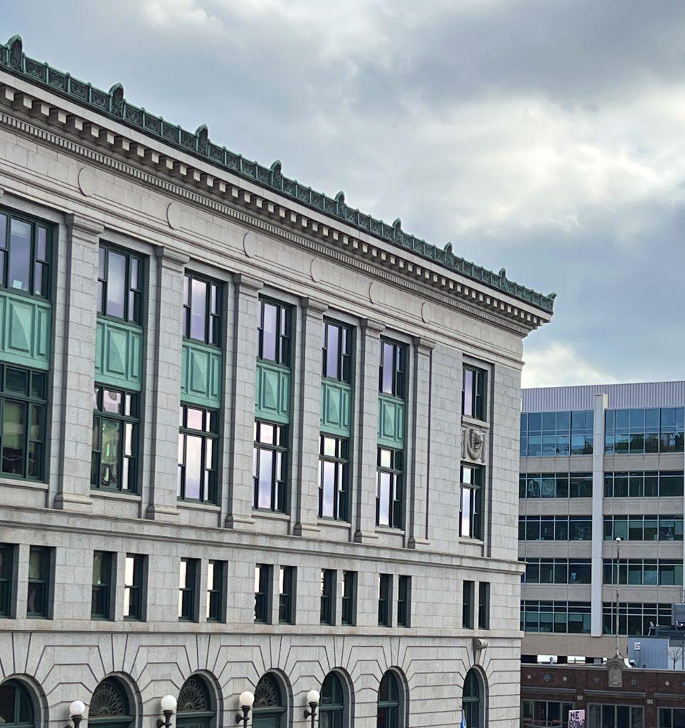 A stone building with tall windows and green decorative panels is shown under a cloudy sky, with a modern glass office building visible in the background.