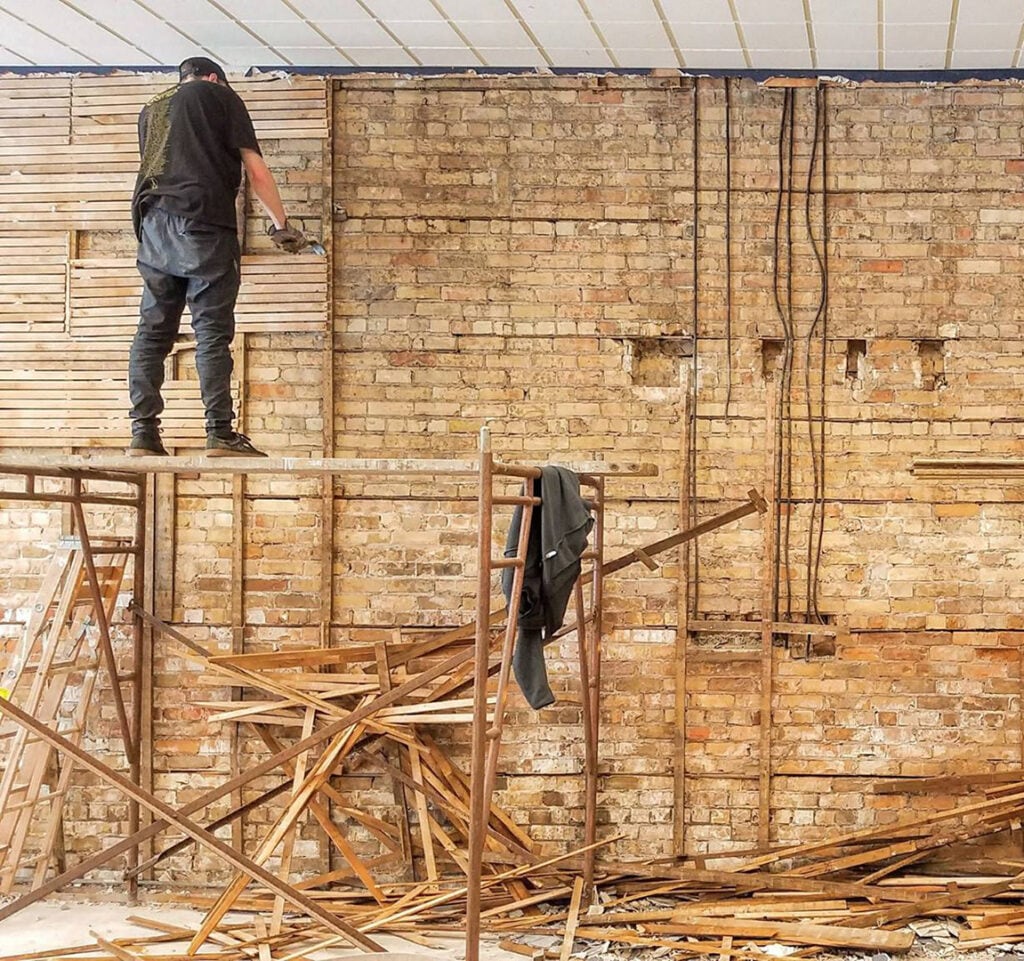 A person stands on scaffolding, removing old wooden slats from a brick wall. Broken wood pieces are scattered on the floor below, and a jacket hangs on the scaffolding. The ceiling panels above are partially visible.