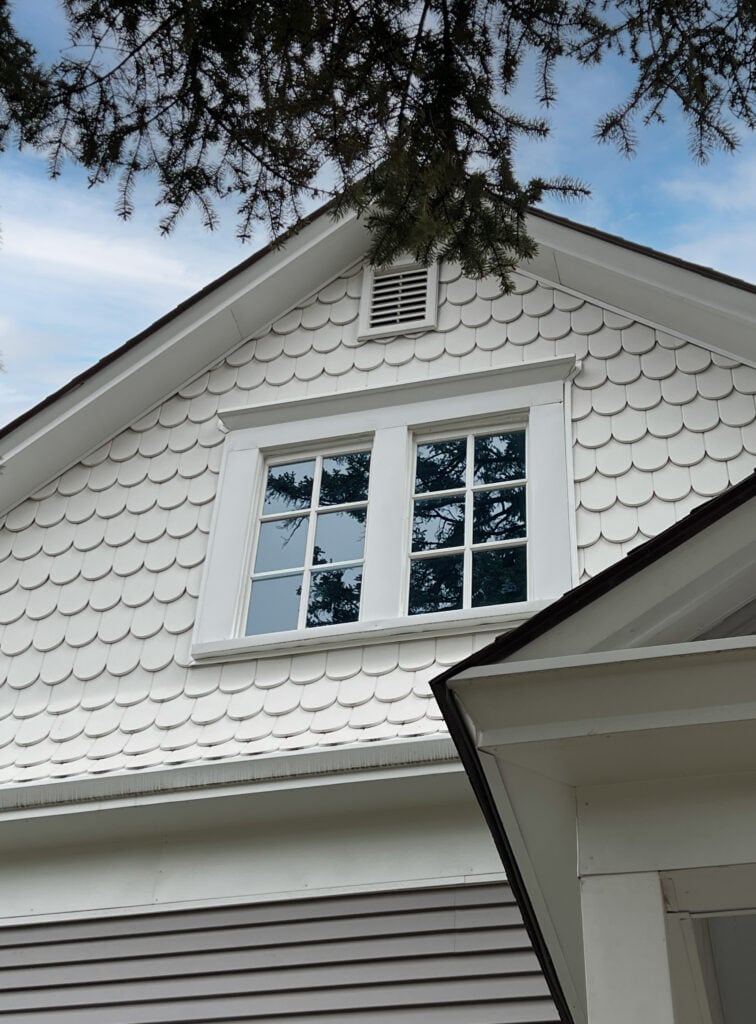 A close-up view of a house gable with white fish scale shingles, a white-framed window, and part of the roof, with tree branches and blue sky above.