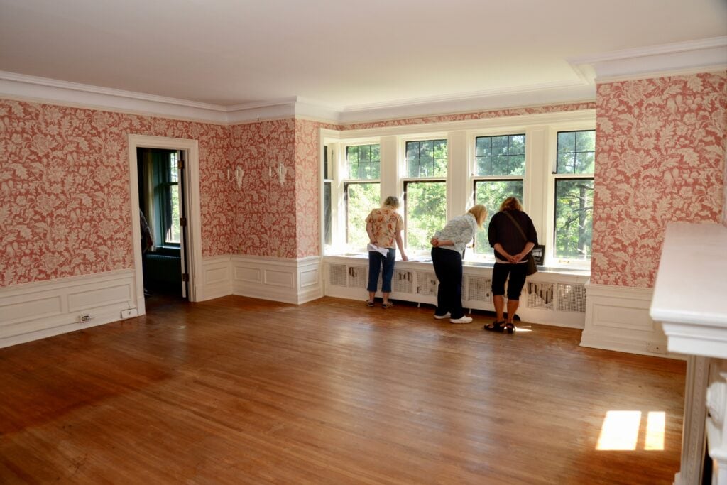 Three people stand by a large window in an empty room with wooden floors, red and white patterned wallpaper, and white trim, looking outside in daylight.