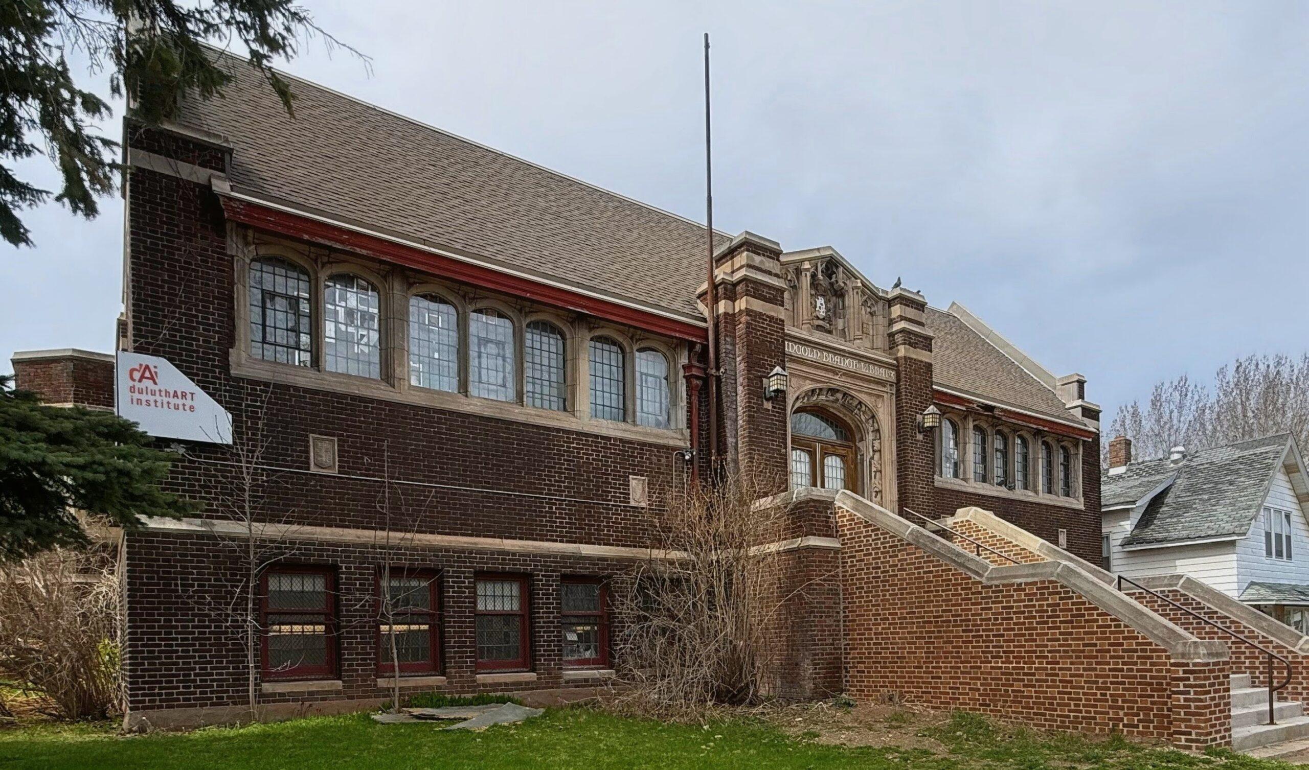 A large, historic brick building with tall windows, a steep roof, and a wide staircase leading to the main entrance; a sign reads “Eastmarket Institute.” Trees and grass surround the structure.