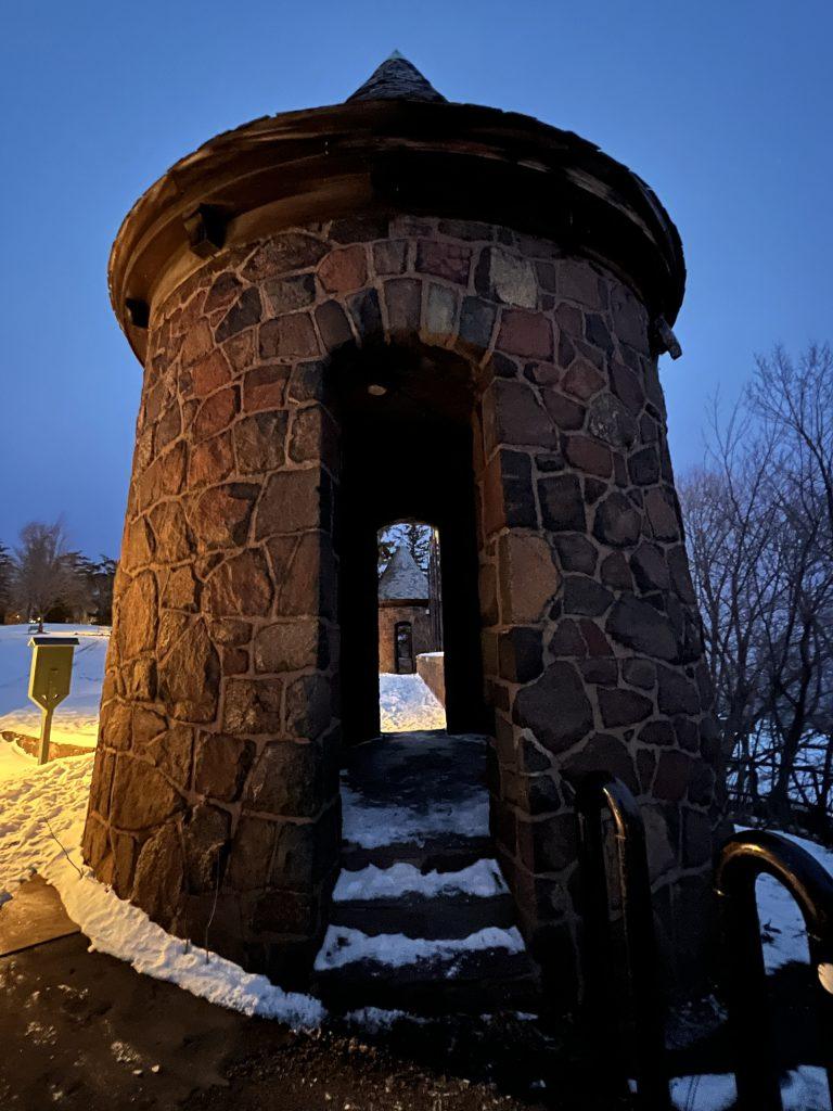A small round stone tower with an arched doorway stands in the snow at dusk. The structure is lit warmly from the left, while the sky and trees in the background appear dark and wintry.