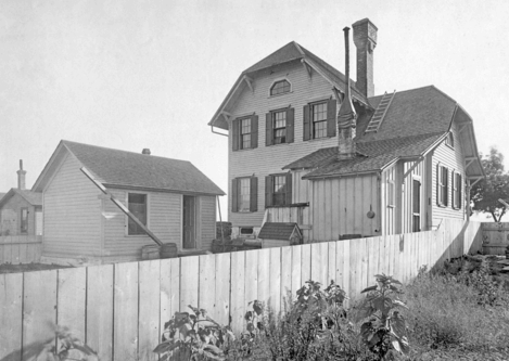 A black and white photo of a two-story house with shutters and a chimney, next to a smaller building, both behind a tall wooden fence with plants in the foreground.