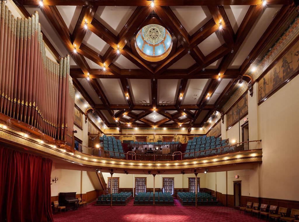 Interior view of a historic theater with ornate wooden ceiling, circular skylight, large pipe organ on the left, red carpet, and rows of blue seats on the main floor and balcony. Warm lighting fills the space.