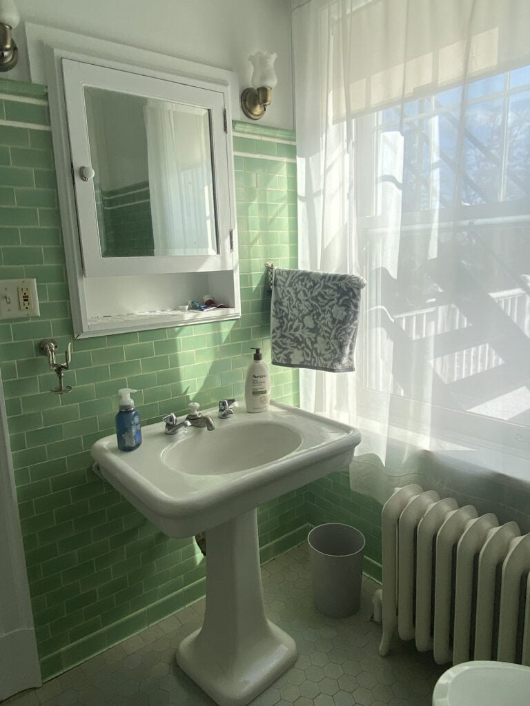 Sunlit bathroom with green tiled walls, a white pedestal sink, liquid soap, a patterned towel hanging, white medicine cabinet, radiator, small trash can, and sheer curtains letting light in through a window.