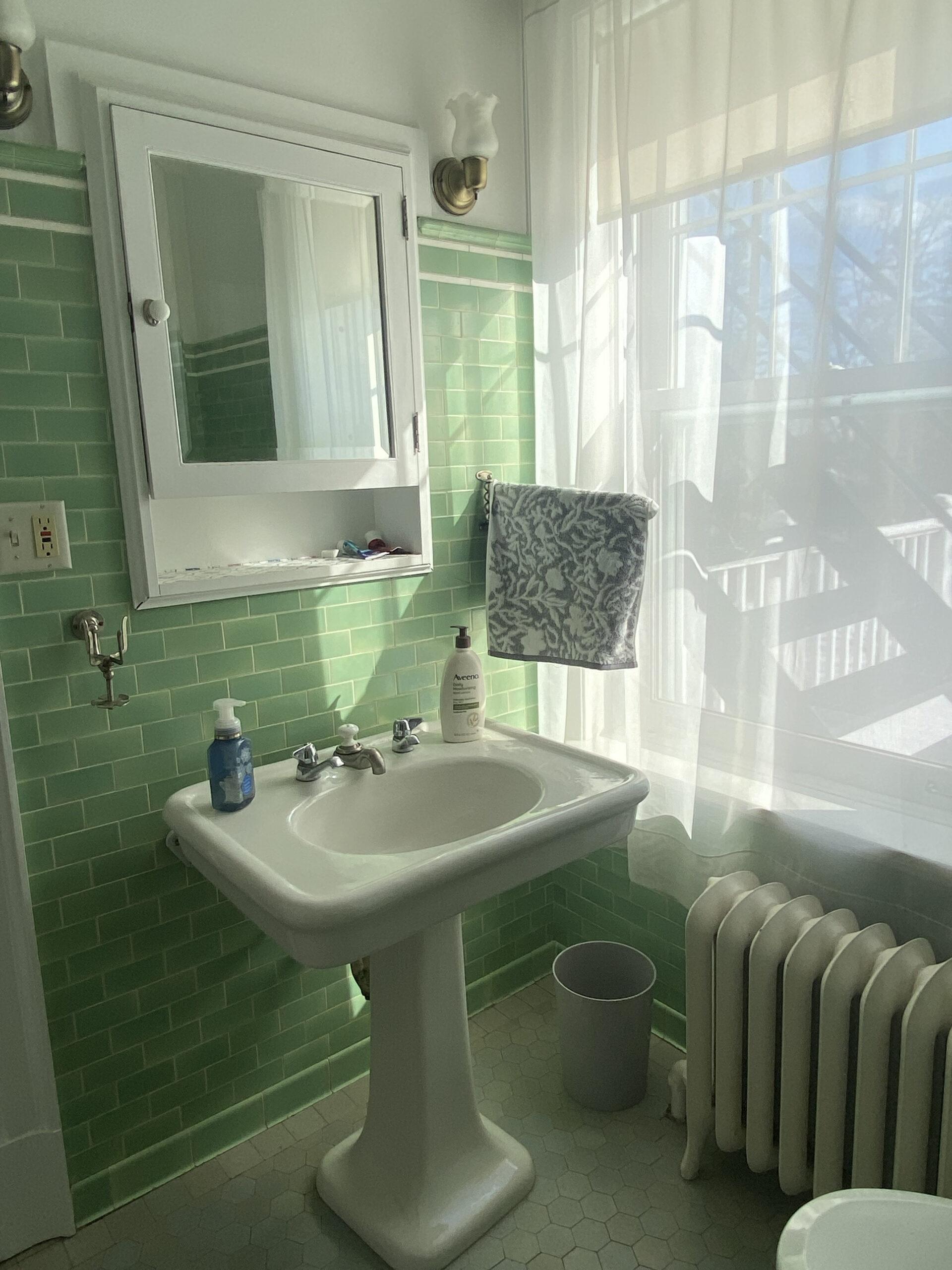 A bathroom with green tile walls, a white pedestal sink, a wall-mounted mirrored cabinet, liquid soap bottles, a towel hanging by the window, a radiator, and sunlight streaming through sheer curtains.