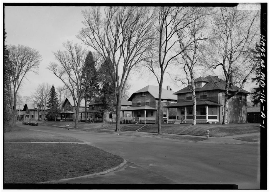 Black and white photo of a quiet suburban street lined with large, early 20th-century houses and leafless trees. The street is empty and lawns are visible in the foreground.