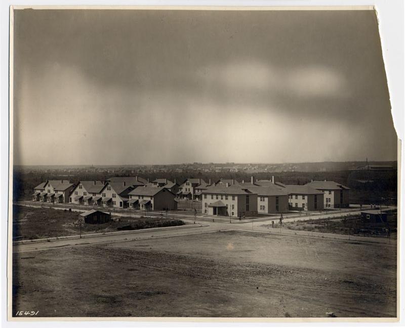 A sepia-toned photograph of a small neighborhood with several two-story residential buildings, arranged along dirt roads, and surrounded by open land under a cloudy sky.