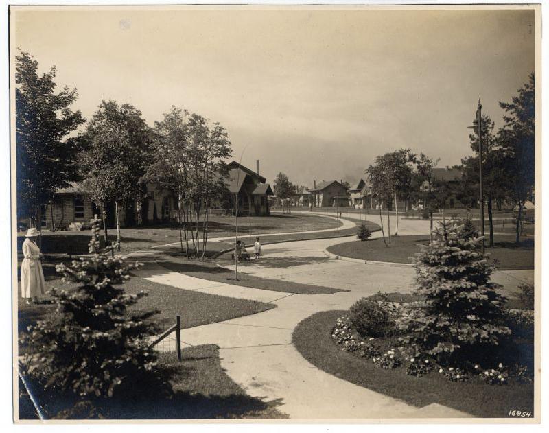 Black-and-white photo of a quiet residential street with curving sidewalks, trees, lawns, and flowerbeds. A woman stands at left, while two children sit on the grass. Houses are visible in the background.