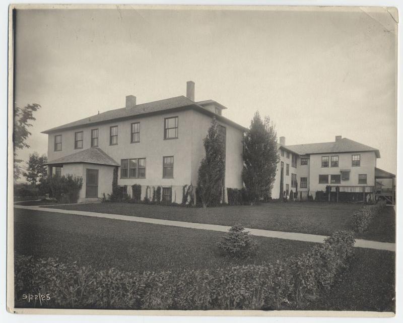Black-and-white photo of two large, two-story stucco houses with rectangular windows, flat roofs, and chimneys, separated by a grassy yard, sidewalks, shrubs, and a few trees.