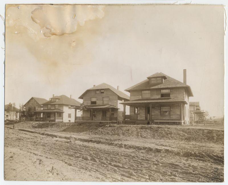 A sepia-toned photo of four early 20th-century houses with porches, standing along a dirt road in a developing neighborhood. The image has visible water stains and rough edges.