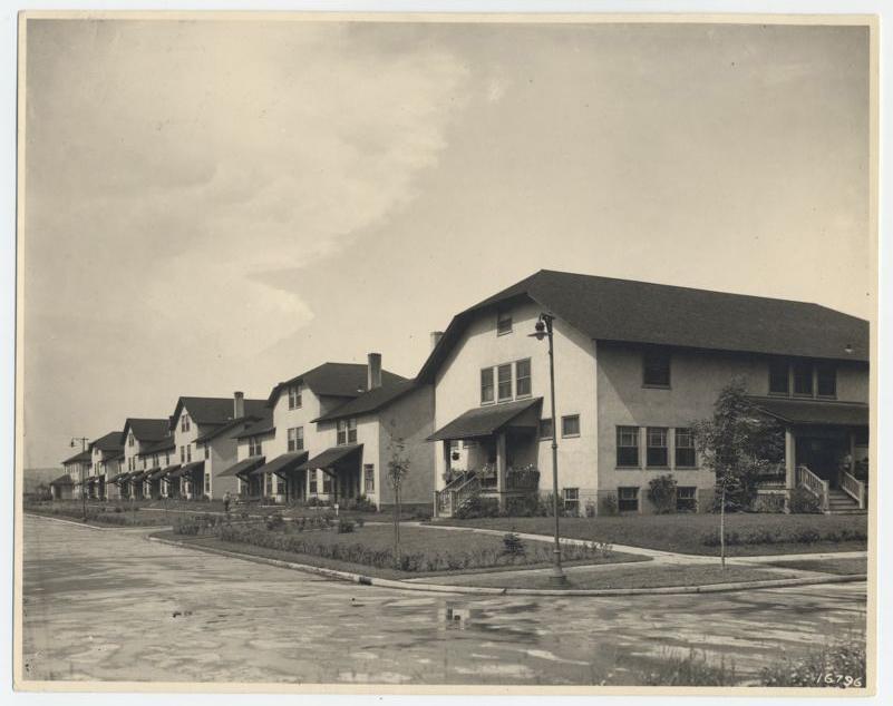 A black and white photo of a row of early 20th-century suburban houses with pitched roofs, lined up along a wet, empty street with sidewalks and small front lawns, under a cloudy sky.