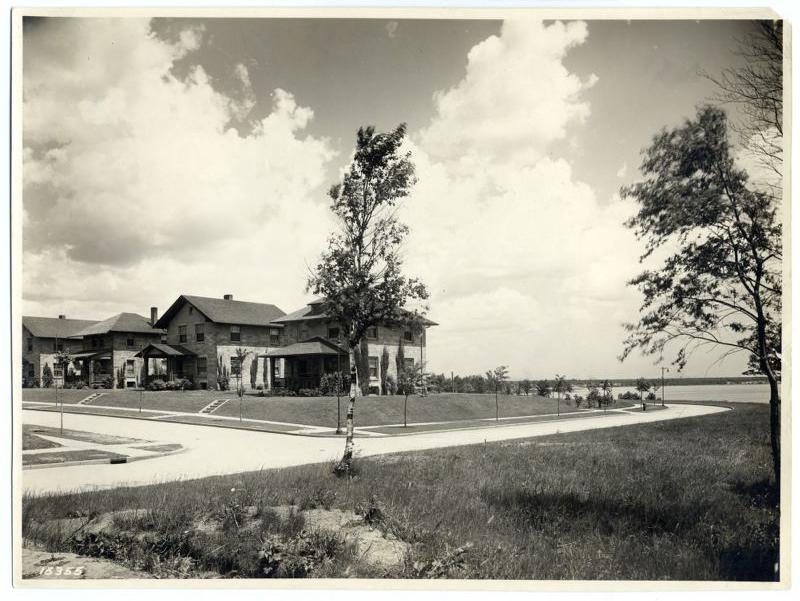 Historic black-and-white photo of large brick houses with porches, a curving sidewalk, grassy lawns, trees, and an open sky with clouds; water is visible in the background.