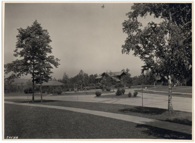 Black and white photo of a park-like area with trees, sidewalks, lawns, and a few small buildings in the background. A single airplane is visible flying high in the sky above the scene.