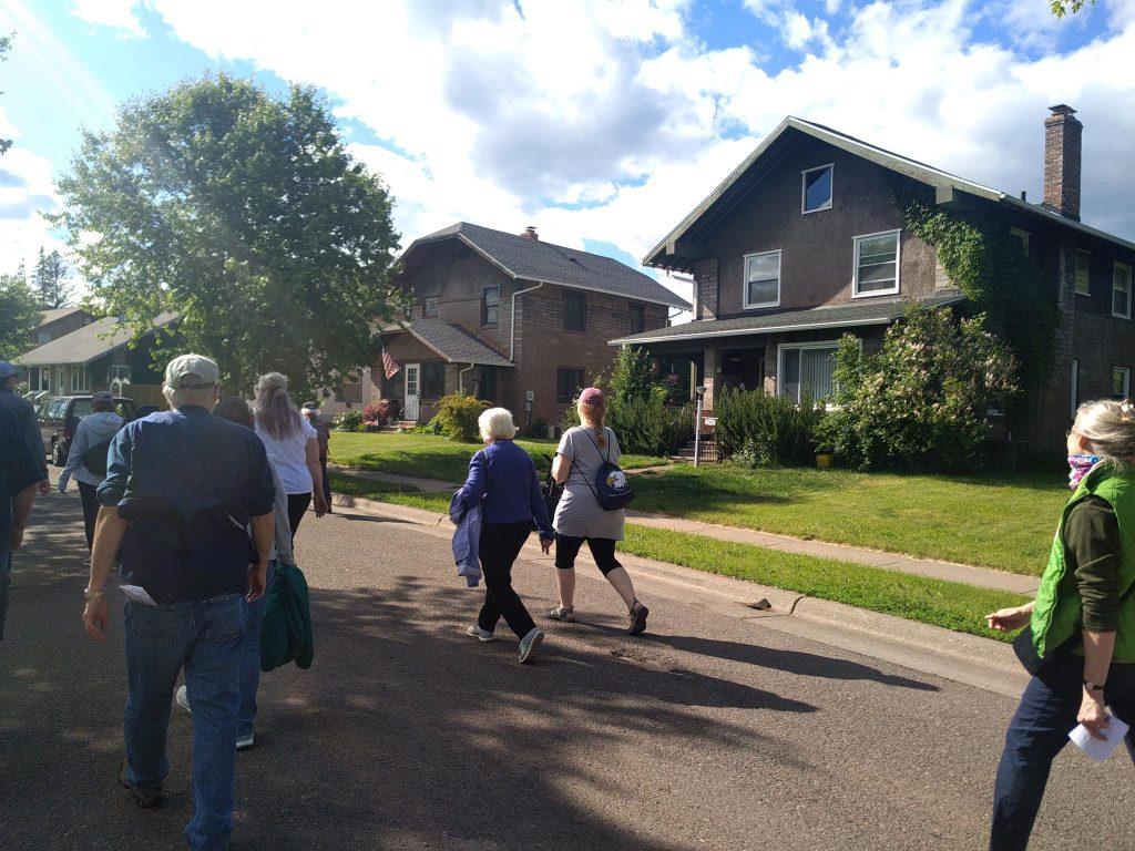 A group of people walk along a sunny residential street lined with trees and two-story houses, some with brick exteriors and greenery. The sky is partly cloudy and everyone appears to be enjoying the day.