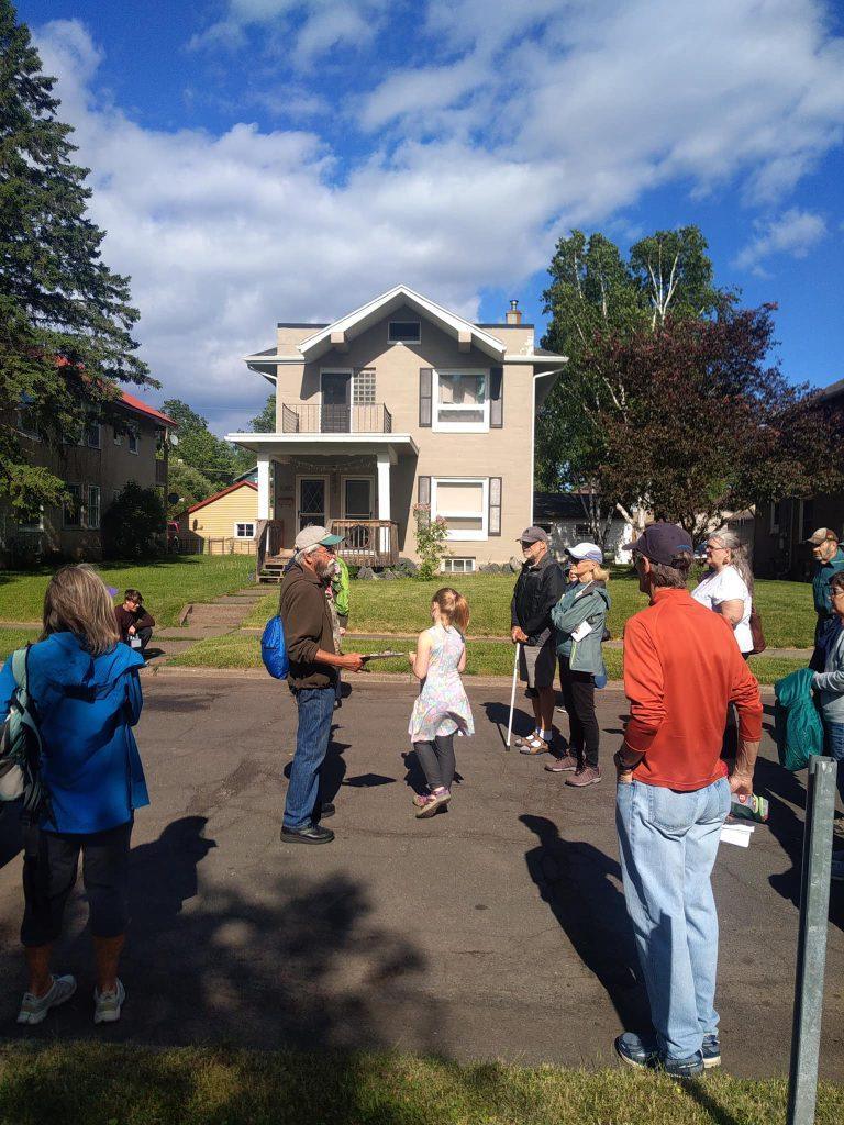A group of people, including adults and children, stand and listen on a sunny street in front of a beige two-story house with trees and blue sky in the background.