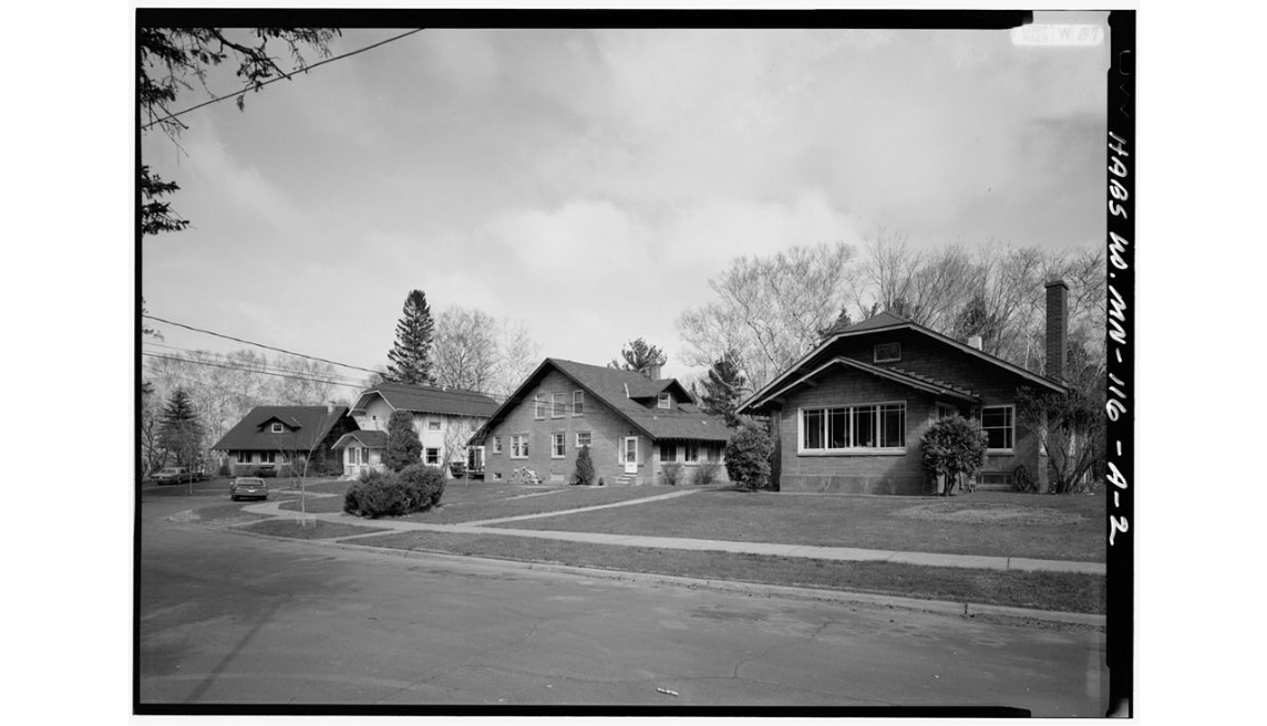 A black-and-white photo of a quiet suburban street with four Craftsman-style houses, large windows, front lawns, leafless trees, and a cloudy sky. A sidewalk runs along the houses, and the street is empty.