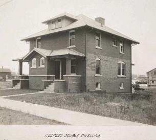 A black and white photo of a two-story brick duplex with covered porches and chimneys, labeled “KEEPERS DOUBLE DWELLING.” The house sits on a grassy lot with a sidewalk and nearby buildings in the background.