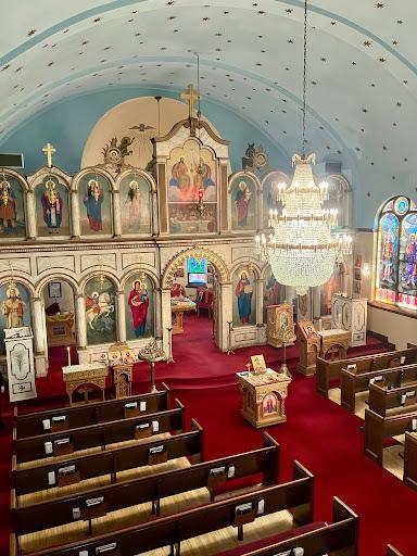 Interior of an ornate church with red carpet, rows of wooden pews, religious icons on the altar, a large chandelier, and colorful stained glass windows. The ceiling is painted light blue with star accents.