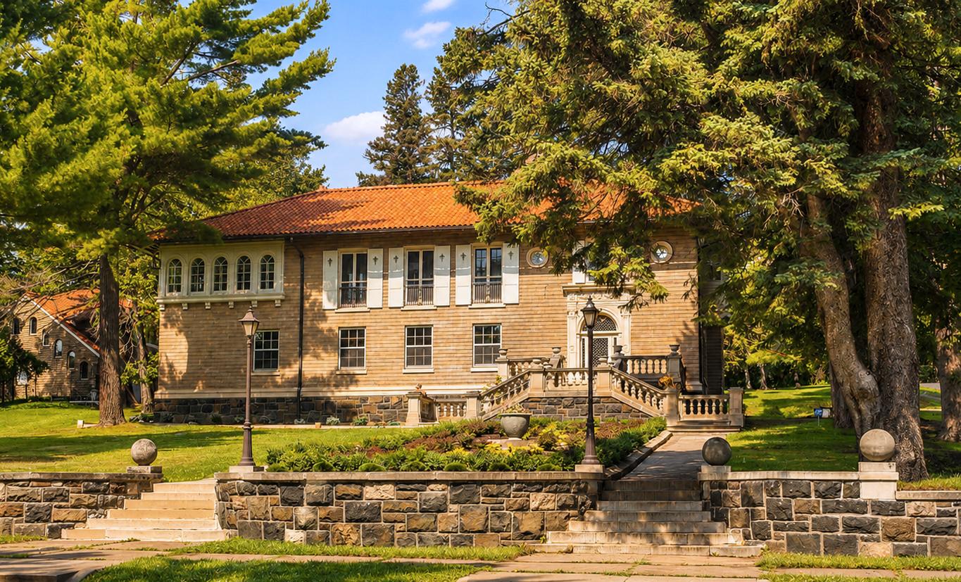 A two-story stone building with white shutters and a red-tiled roof sits behind a stone wall and staircase, surrounded by tall green trees and a grassy lawn under a blue sky.