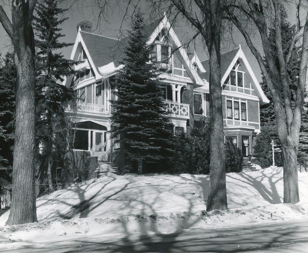 A large, two-story Victorian-style house with gabled roofs and many windows sits behind snow-covered ground and tall leafless trees on a sunny winter day. Evergreen trees partially obscure the front of the house.