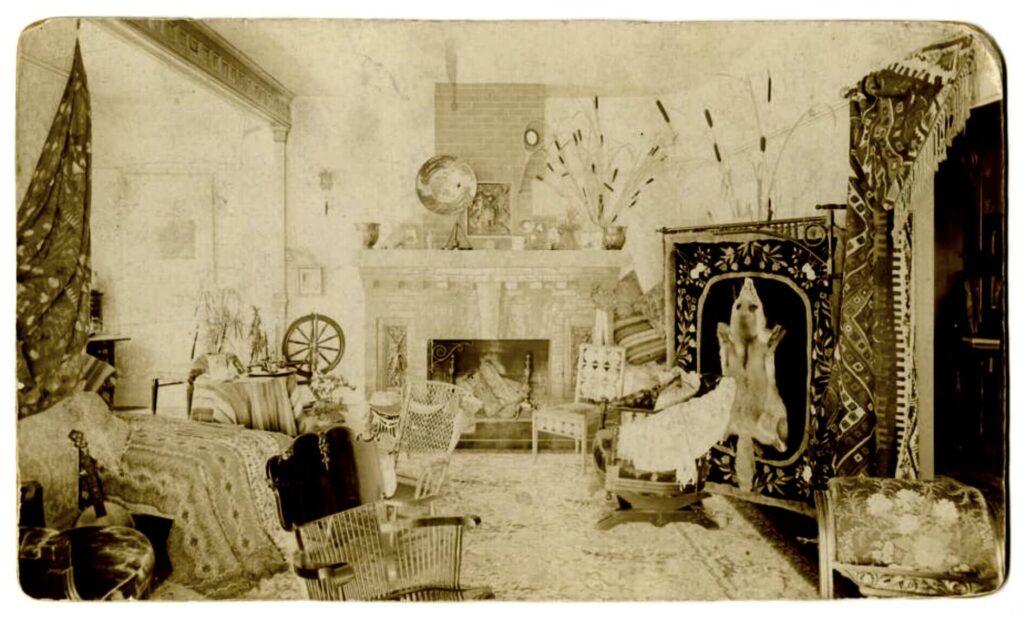Sepia-toned photo of a Victorian-era living room with wicker chairs, ornate rugs, a fireplace, spinning wheel, globe, decorative tapestry featuring a bear, and plants scattered around the cozy, eclectic space.