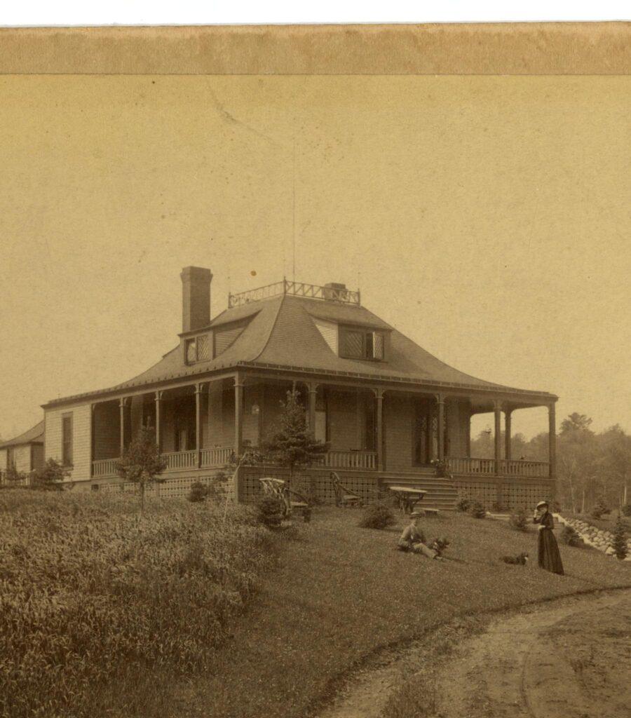 Sepia-toned photo of a large, Victorian-style house with a wraparound porch, surrounded by shrubs and trees. A woman in a long dress stands on the lawn, with a dog laying nearby and another person sitting on the grass.