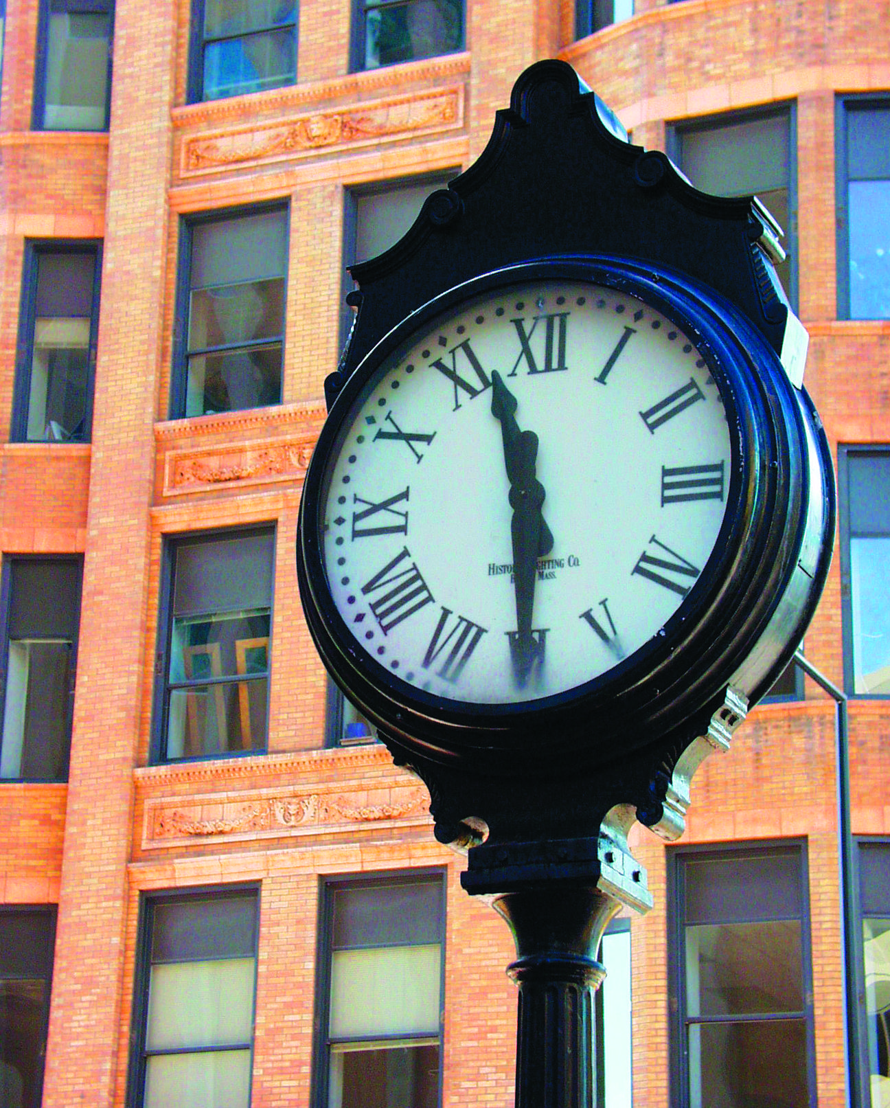 A black vintage street clock with Roman numerals shows the time as 11:58, standing in front of a brick building with large windows.