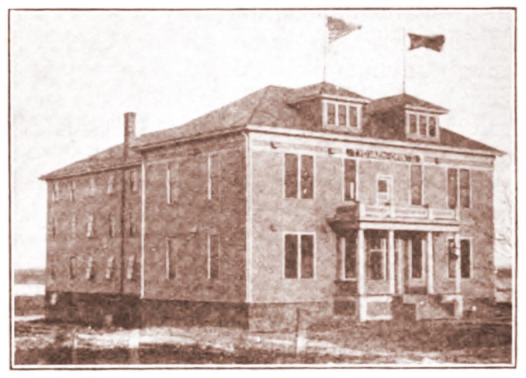 A sepia-toned photo of a two-story brick building with a small porch and two flags flying on the roof. The structure has several large windows and sits on an open plot of land.