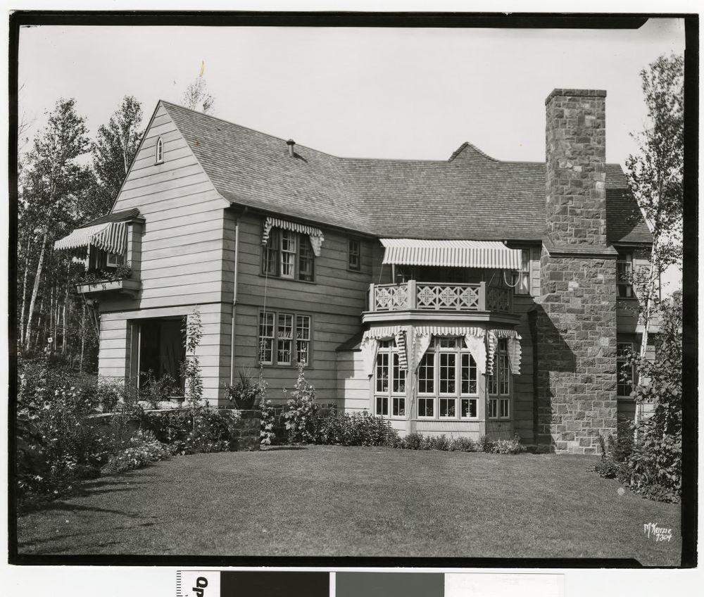 Black and white photo of a two-story house with wood siding, large stone chimney, striped window awnings, a small balcony, and a bay window, surrounded by trees and a manicured lawn with flower beds.