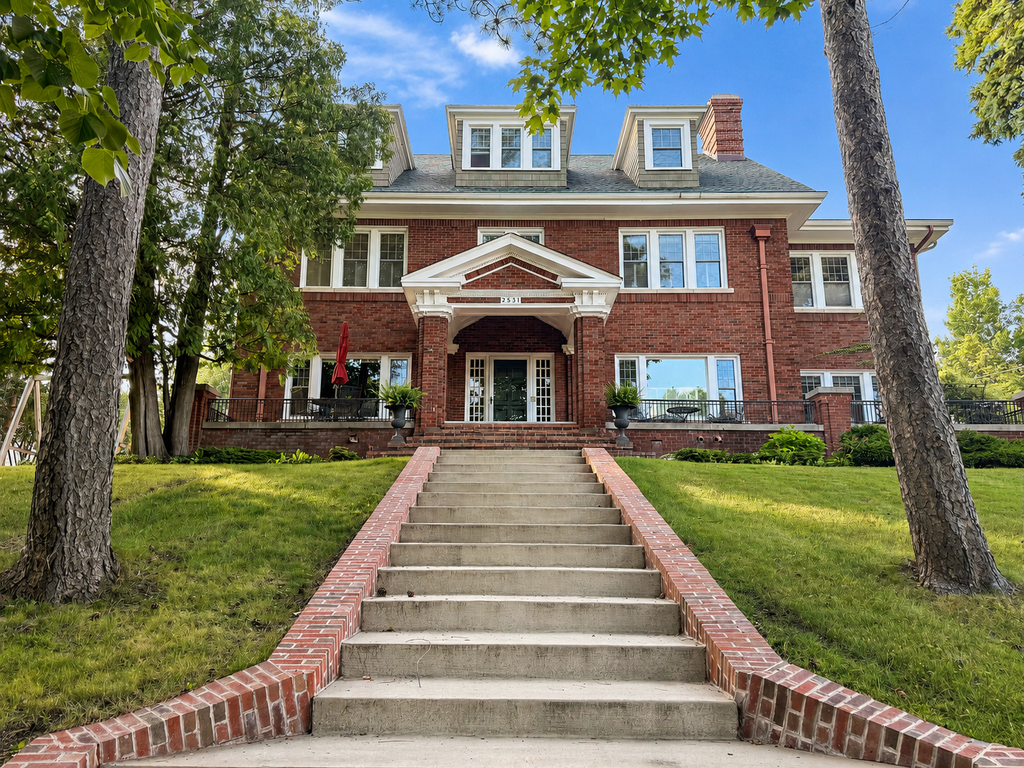 A wide brick staircase leads up a sloped lawn to a large three-story red-brick house with white trim, front porch, and tall trees surrounding the property on a sunny day.
