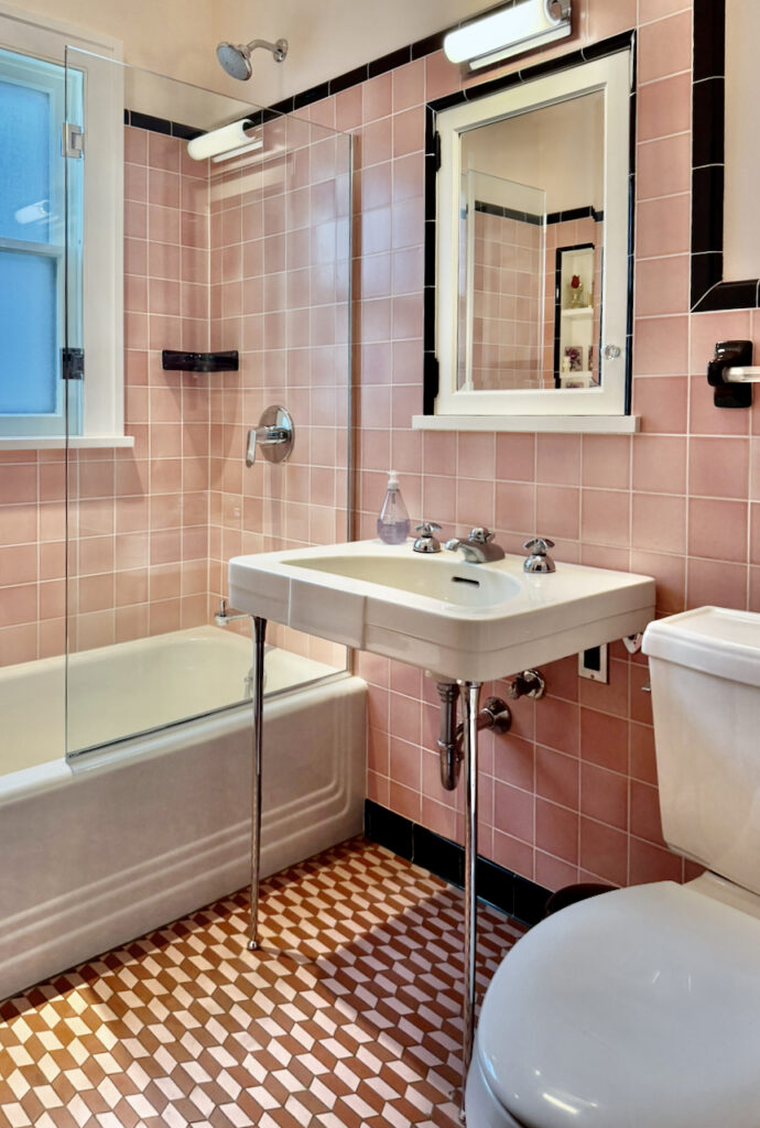 A bathroom with pink wall tiles, a white bathtub with glass door, a white pedestal sink, a toilet, and a mirror above the sink. The floor has a pink and white checkered pattern. Lights are above the mirror and shower.