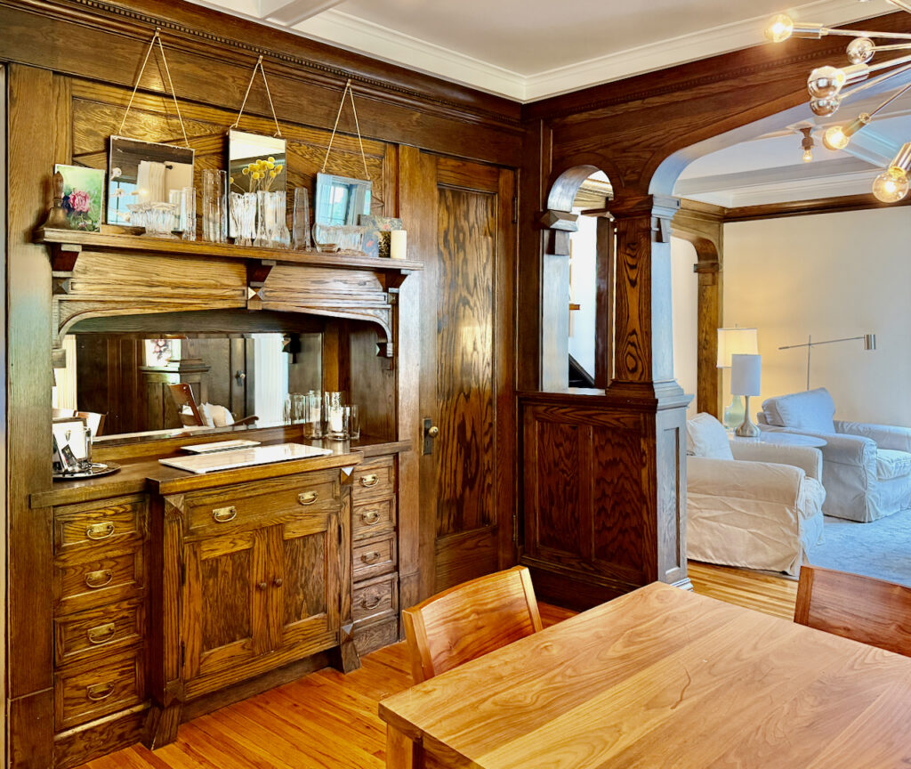 A dining room with ornate dark wood built-ins, drawers, and mirror, leading into a living room with white sofas, light wood floors, and modern lighting. Artwork and decor are placed on the cabinetry and table.