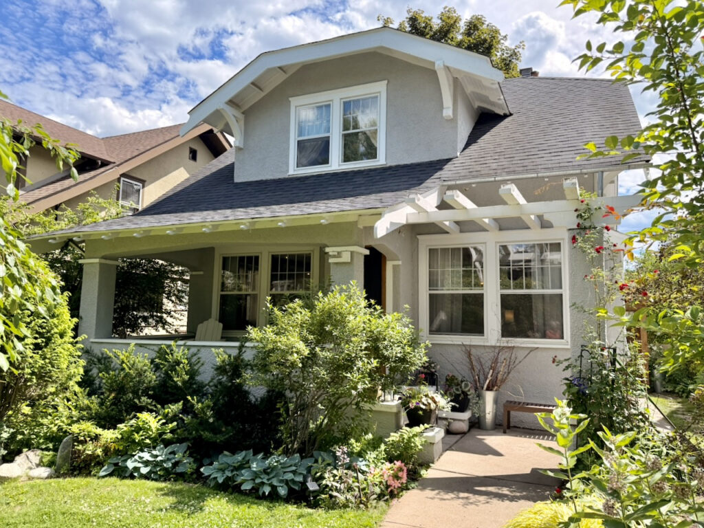 A charming gray bungalow with white trim, a covered front porch, lush green plants, and a pergola. The yard is filled with flowers and greenery under a partly cloudy sky.