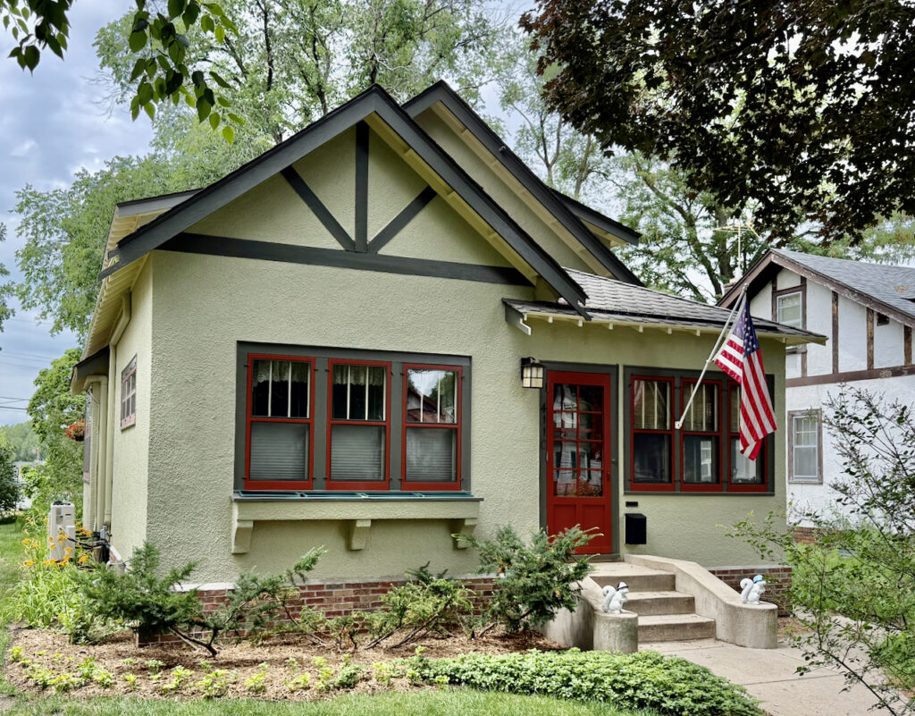 A small light green house with red-trimmed windows and door, a front porch, an American flag, flower beds, and bushes in front, surrounded by trees and neighboring houses.