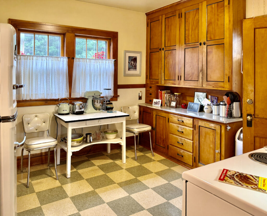 Bright vintage kitchen with wood cabinets, white appliances, retro chairs, and checkered floor. A table holds a mixer and tea kettle under curtained windows. Various kitchen tools and framed photos are on the countertop.