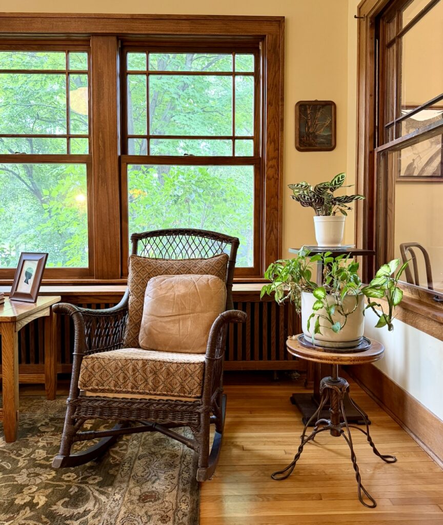A cozy corner with a wicker rocking chair, tan pillow, side table with potted plants, wooden windows letting in green outdoor light, and a framed photo on a small table.