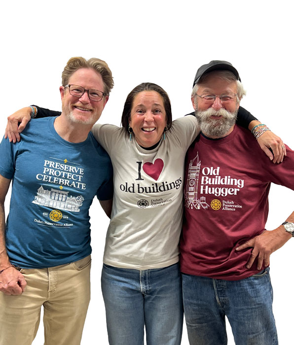 Three people with arms around each other, wearing shirts that say “Preserve Protect Celebrate,” “I love old buildings,” and “Old Building Hugger,” all featuring Duluth Preservation Alliance branding.